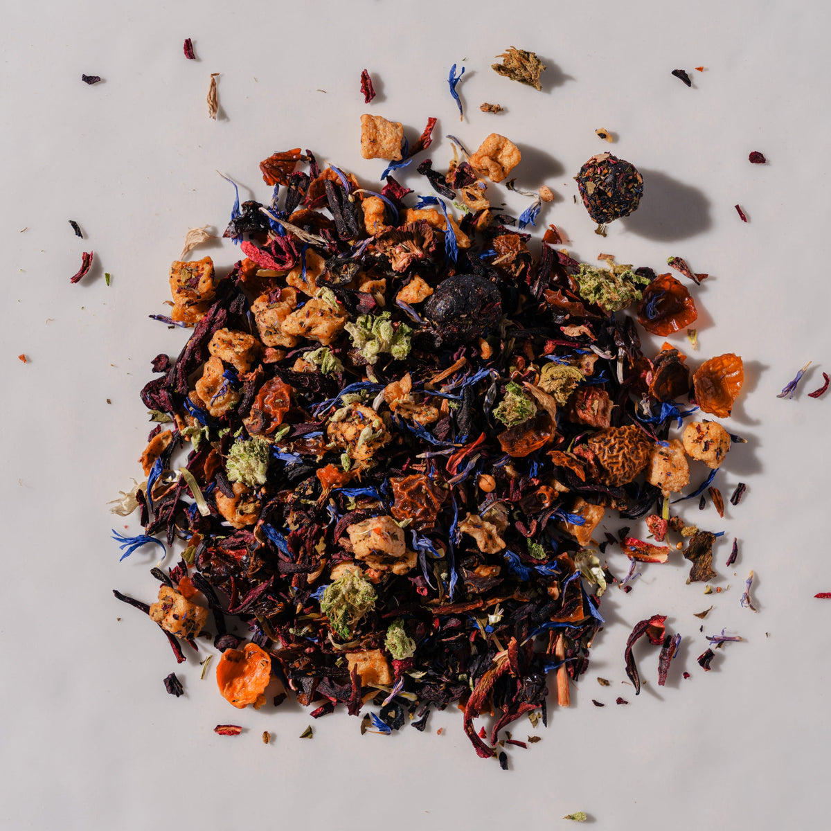 A pile of loose-leaf herbal tea blend on a light gray background, featuring dried flowers, citrus slices, and dark tea leaves in orange, red, blue, green, and brown tones.
