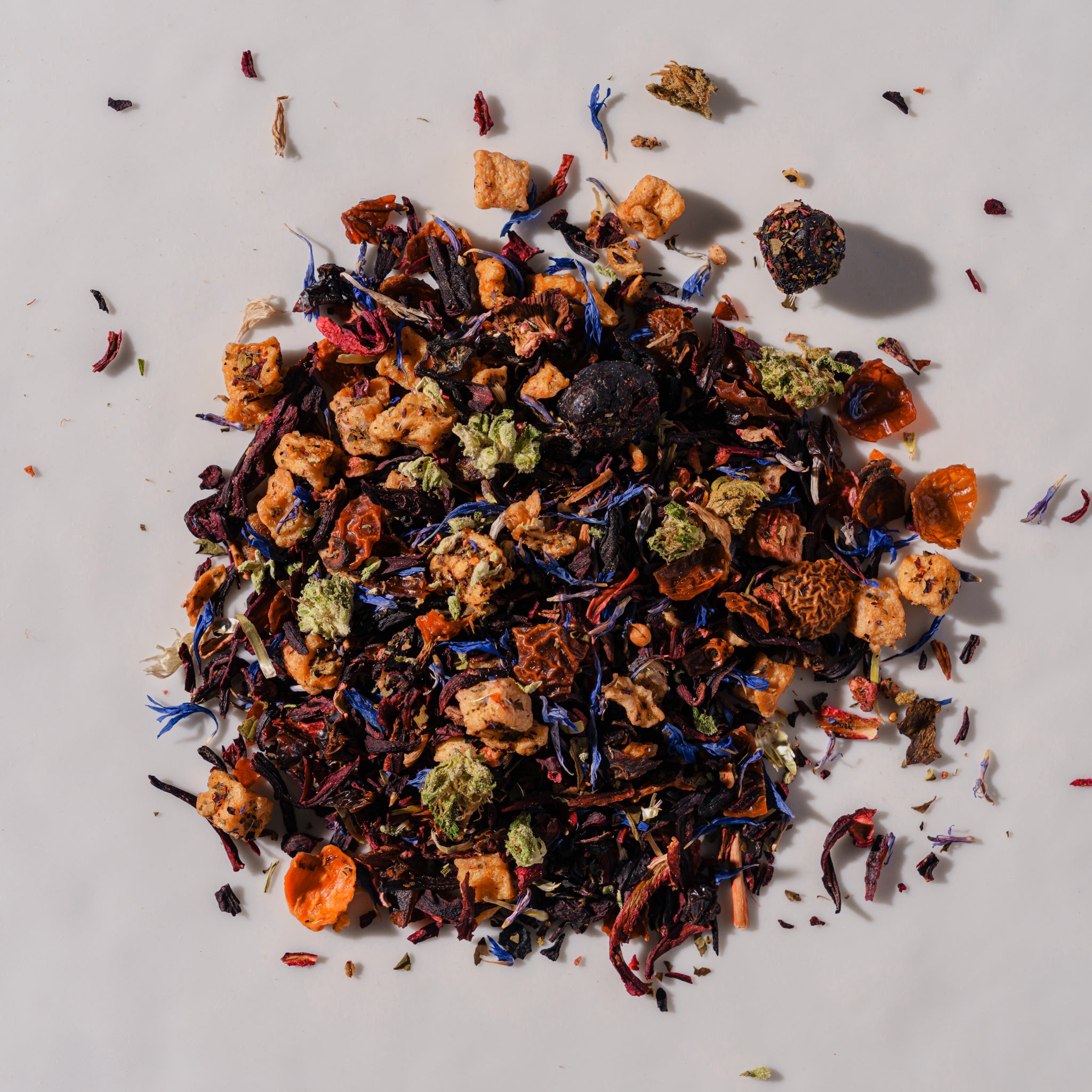 A pile of loose-leaf herbal tea blend on a light gray background, featuring dried flowers, citrus slices, and dark tea leaves in orange, red, blue, green, and brown tones.