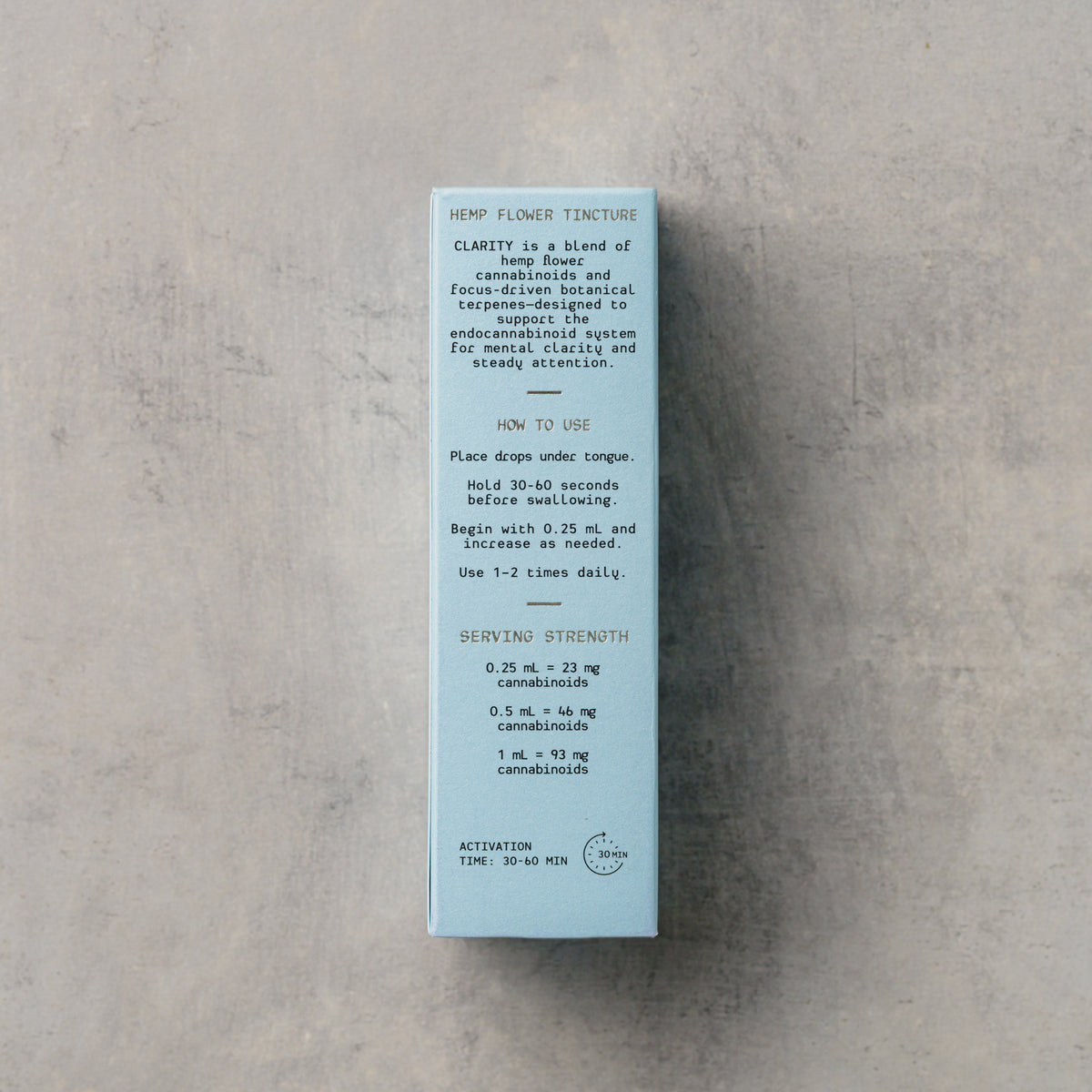 A tall, narrow light blue box with black text, photographed from above on a light gray textured surface. The box appears to be the packaging for a liquid product, with the back side visible.